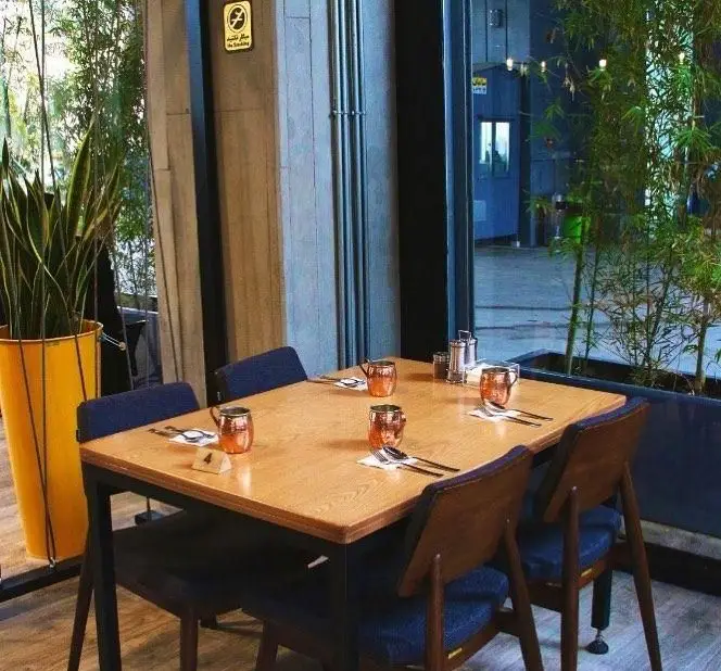 فضای داخلی دنج و مدرن رستوران/کافه م س ی ن با میز چوبی چهار نفره، لیوان‌های مسی، صندلی‌های آبی و گیاهان طبیعی در گلدان.