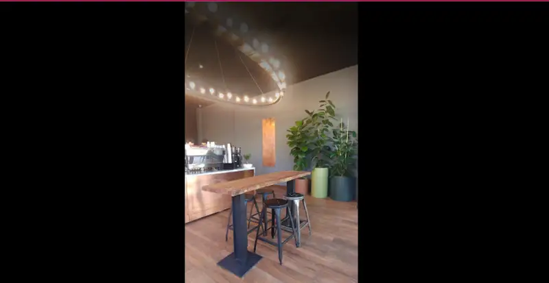 فضای داخلی شیک و مدرن کافه لمیز شهرک غرب؛ میز چوبی بلند، صندلی‌های کانتر، لوستر دایره‌ای و گیاهان سبز طبیعی.