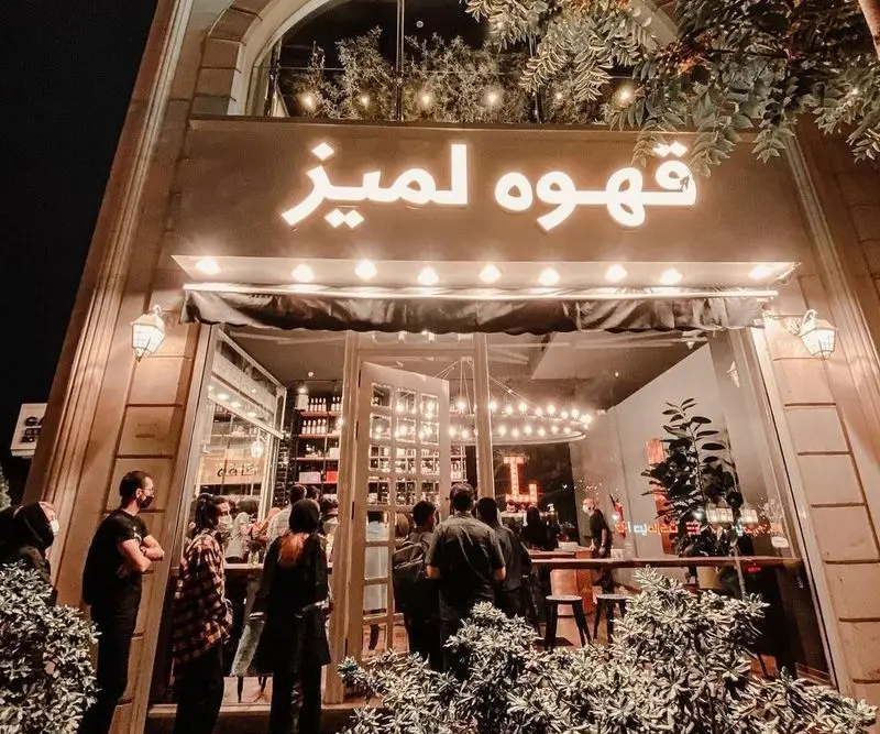 نمای شب کافه لمیز شهرک غرب با تابلوی "قهوه لمیز" و مشتریان در حال ورود به فضایی دلپذیر برای تجربه ای لذت بخش.