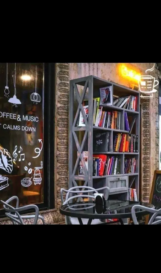 فضای دنج کافه لید با قفسه کتاب پر از کتاب، میز و صندلی، نمادهای قهوه و موسیقی روی پنجره و نور نئون. ایده آل برای مطالعه و آرامش.