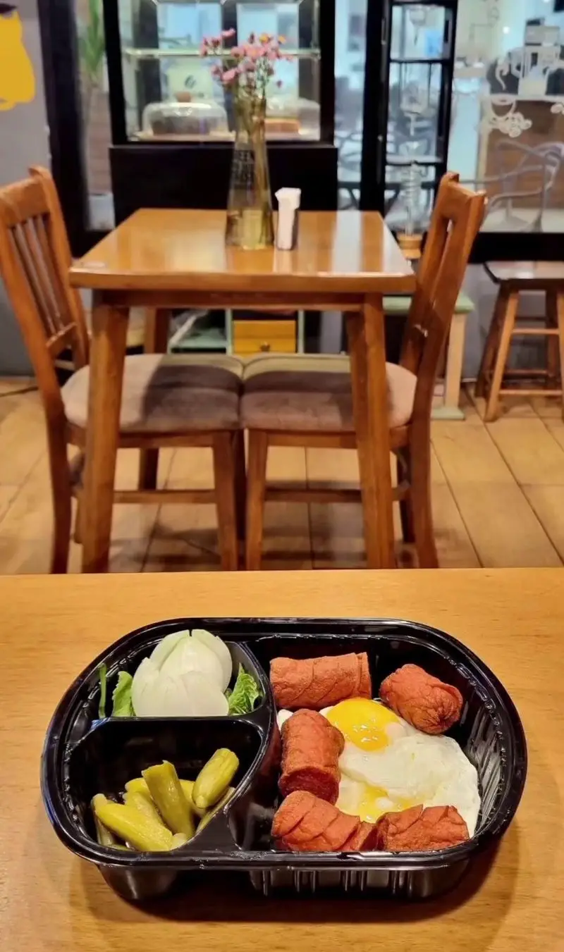 وعده صبحانه یا ناهار کافه لید شامل سوسیس، تخم مرغ، خیارشور و سالاد، آماده برای دلیوری یا سرو در فضای دنج کافه با میز چوبی.