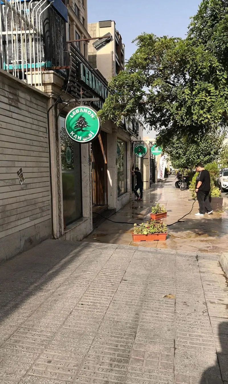 نمای بیرونی کافه لبنان (Lebanon Cafe) با تابلوهای سبز رنگ و نماد درخت سرو، پیاده‌رو تمیز، و محیطی دلنشین برای قهوه و دسر.