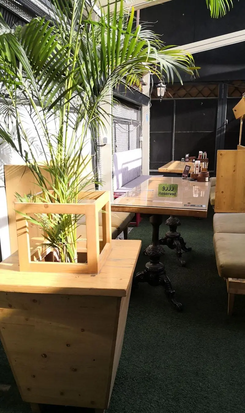 نمای دنج کافه لبنان با فضای سبز، میزهای چوبی و نشیمن راحت، مکانی ایده‌آل برای لذت بردن از لحظات آرام. یک میز رزرو شده نیز دیده می‌شود.