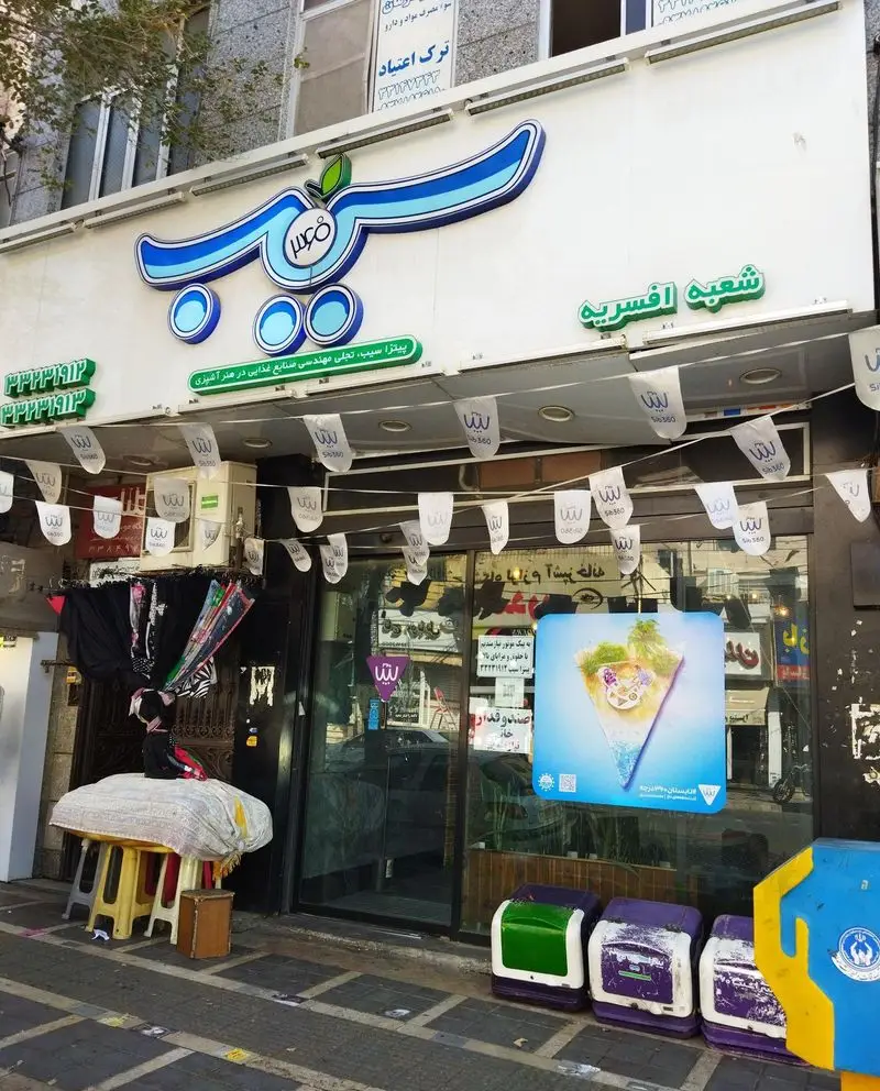 تصویر نمای بیرونی شعبه افسریه پیتزا سیب ۳۶۰، فست فود معروف در تهران، با تابلوی بزرگ و جذاب، ورودی شیشه‌ای و تبلیغات روی بنرها.