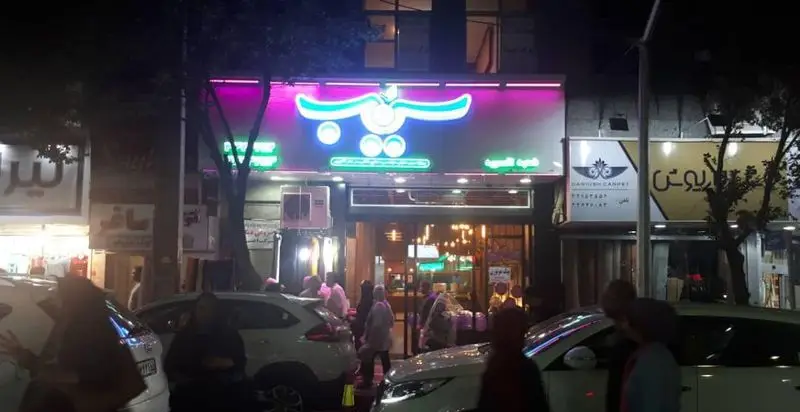 تصویری شبانه از نمای بیرونی رستوران پیتزا سیب ۳۶۰ با تابلوی نئونی جذاب و شلوغی مشتریان و خودروها در خیابان، یک فضای پویا.