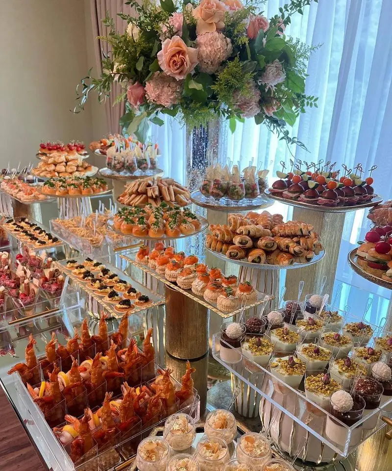 میز بوفه مجلل با گل آرایی زیبا از بالونیا، شامل انواع فینگرفود، دسر و غذاهای مهمانی. طراحی شیک و حرفه ای مراسم