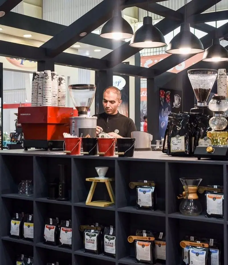 باریستا در کافه رئیس مشغول آماده‌سازی قهوه، در حالی که دستگاه اسپرسو، دانه و بلندهای قهوه و تجهیزات حرفه‌ای به نمایش گذاشته شده‌اند.