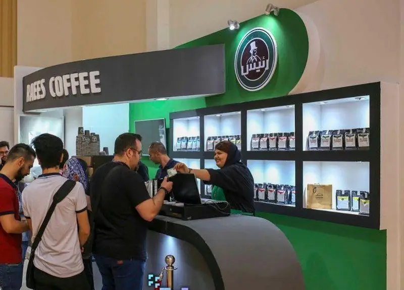 نمایی پویا از کافه رئیس با مشتریانی که از پیشخوان خرید قهوه می‌کنند. قفسه‌های نورانی با بسته‌های متنوع قهوه، دکوراسیون مدرن سبز و لوگوی برجسته.