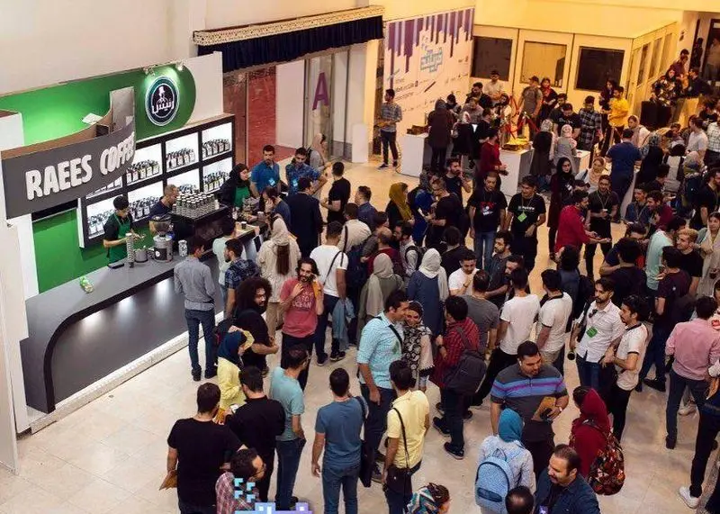 غرفه شلوغ کافه رئیس در نمایشگاهی پر جنب و جوش، با بارسیتاهای فعال در سرو قهوه و مشتریان مشتاق در حال خرید محصولات متنوع.