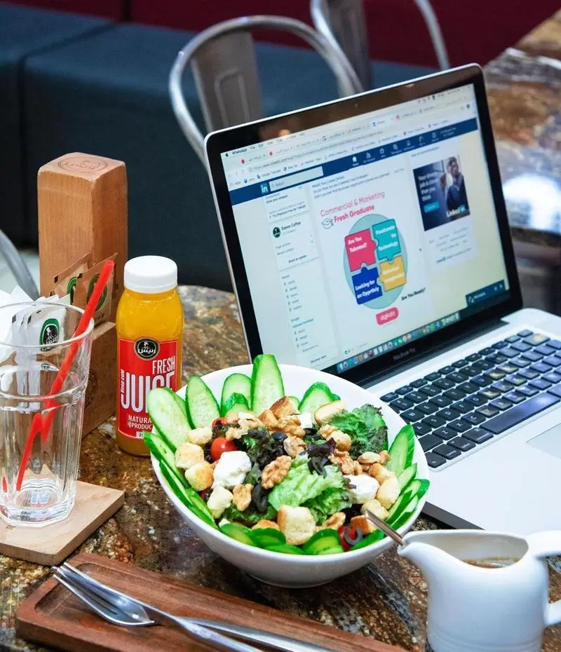 فضای کافه رئیس برای کار و مطالعه: لپ تاپ، سالاد سالم و مغذی، آبمیوه تازه طبیعی رئیس، لیوان آب و سس سالاد. محیطی ایده آل برای دورکاری.