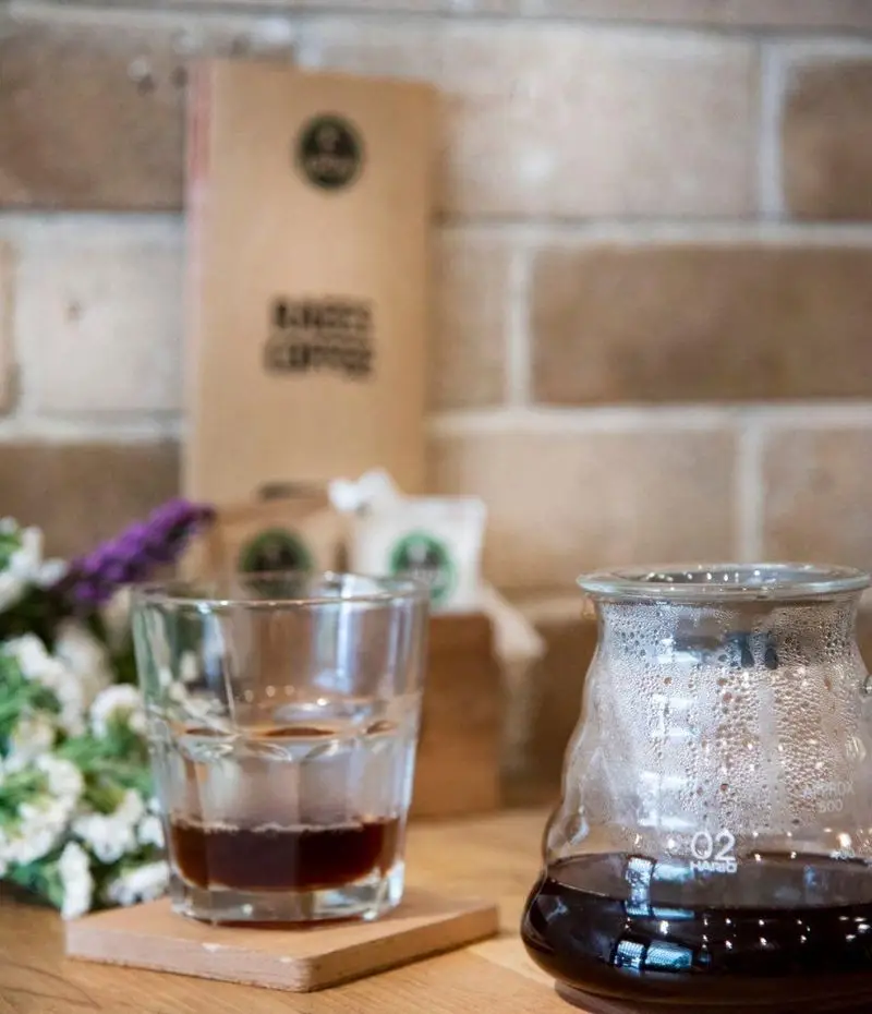 تصویری جذاب از یک میز چوبی در کافه رئیس با فنجان قهوه، قهوه ساز شیشه‌ای و بسته بندی‌های قهوه مخصوص. لذت قهوه‌ای باکیفیت را تجربه کنید.