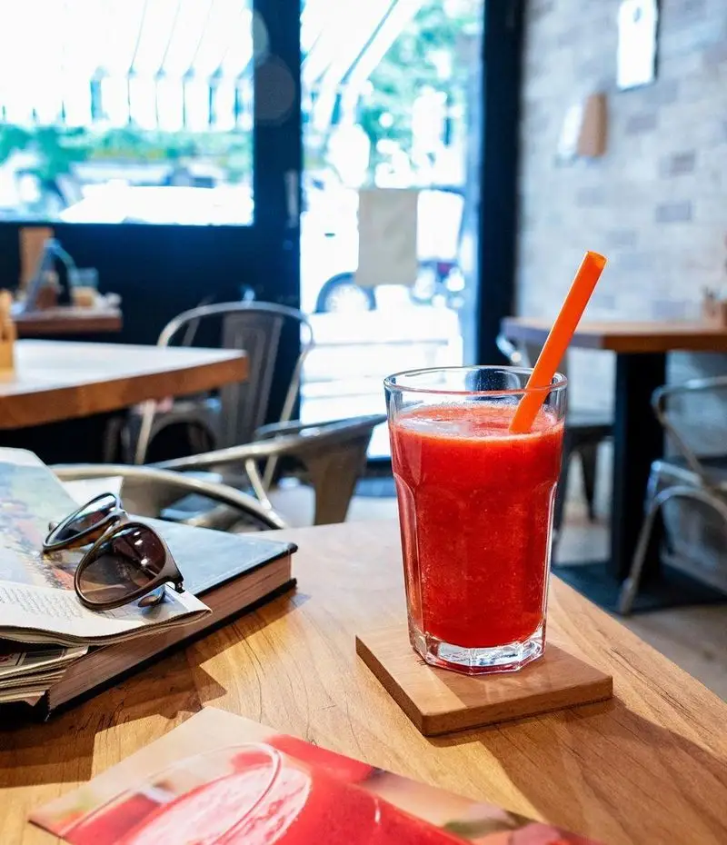 نوشیدنی خنک و گوارا در کافه رئیس: اسموتی میوه قرمز با نی نارنجی روی میز چوبی، همراه با کتاب و عینک آفتابی در فضایی آرام و دلنشین.