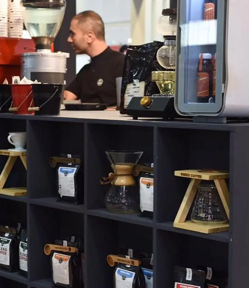 کافه رئیس: باریستا پشت پیشخوان قهوه ساز، شلف‌های مملو از دانه قهوه، محصولات و تجهیزات دم آوری قهوه شیک.