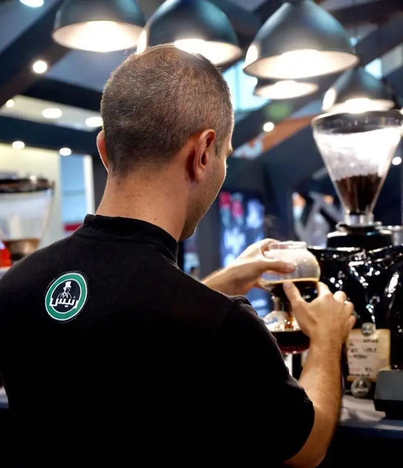 باریستای حرفه ای کافه رئیس از پشت در حال آماده سازی قهوه با دقت، لوگوی معروف کافه بر روی یونیفرم مشکی او به وضوح دیده می‌شود.