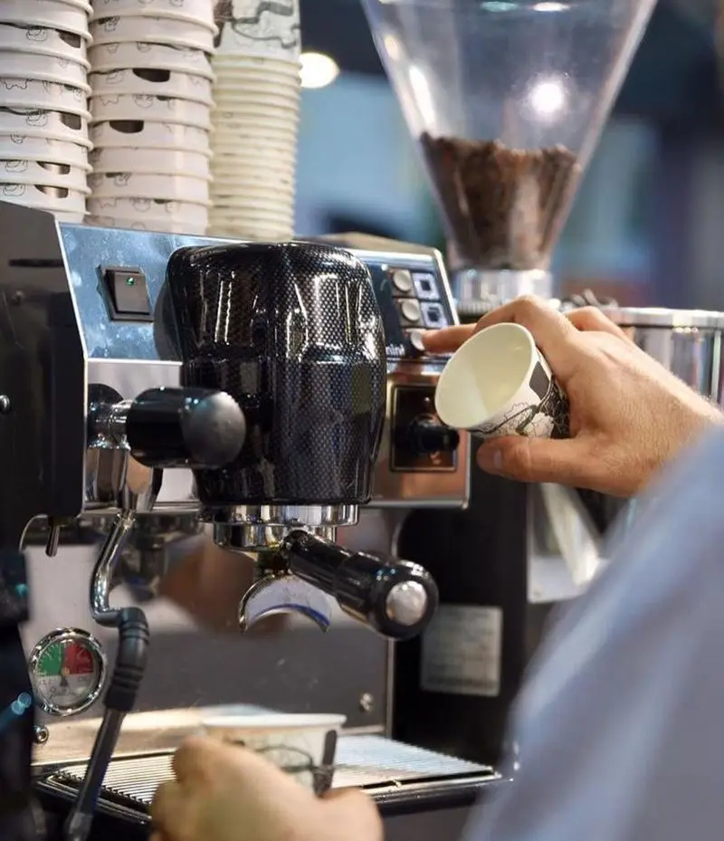باریستا در کافه رئیس، مشغول آماده‌سازی قهوه اسپرسو با دستگاه حرفه‌ای، فنجان‌های کاغذی آماده برای سرو.