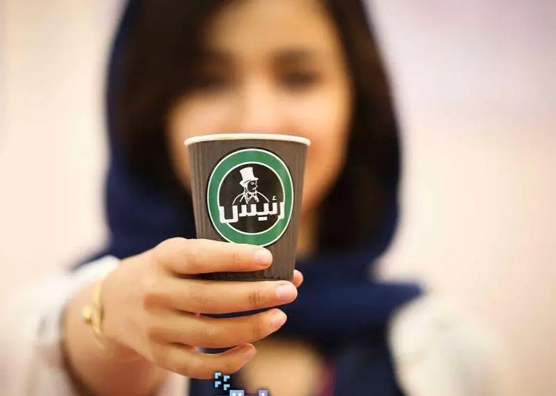 زن جوانی با حجاب در حال نگه داشتن یک فنجان قهوه از کافه رئیس است. لوگوی سبز رنگ "رئیس" با تصویر مرد کلاهدار روی کاپ تیره خودنمایی می‌کند.