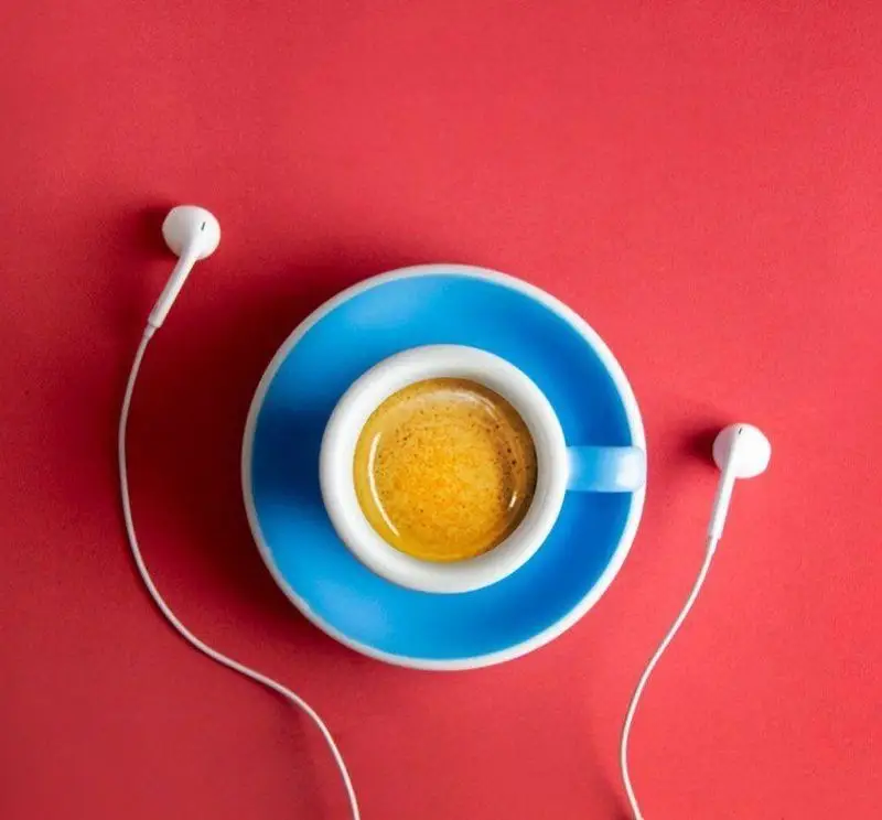 یک فنجان اسپرسو با نعلبکی آبی و هدفون‌های سفید روی پس‌زمینه قرمز، لحظات آرامش‌بخش کافه رئیس را نشان می‌دهد.