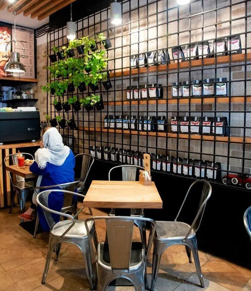 کافه رئیس: نمای داخلی مدرن با قفسه‌های نمایش قهوه، بسته‌بندی‌های متنوع و فضای دلنشین برای مشتریان با گیاهان سبز.