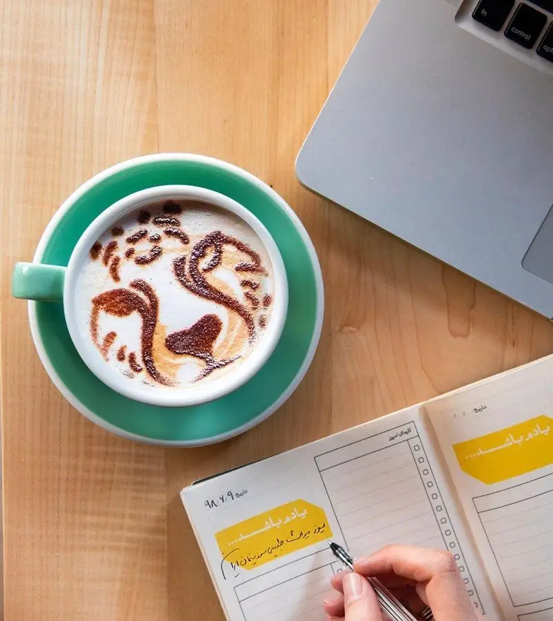 قهوه لاته آرت طرح ببر در فنجان فیروزه‌ای روی میز چوبی، همراه با لپ تاپ و دفتر یادداشت در کافه رئیس برای برنامه‌ریزی روزانه.