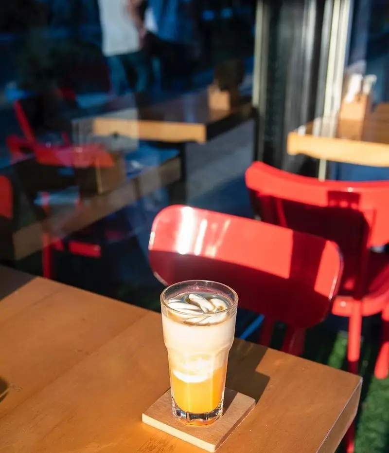 نوشیدنی آیس لاته خنک و خوشمزه در کافه رئیس، روی میز چوبی روشن با انعکاس نور خورشید، صندلی های قرمز دکوراسیون دلنشین.