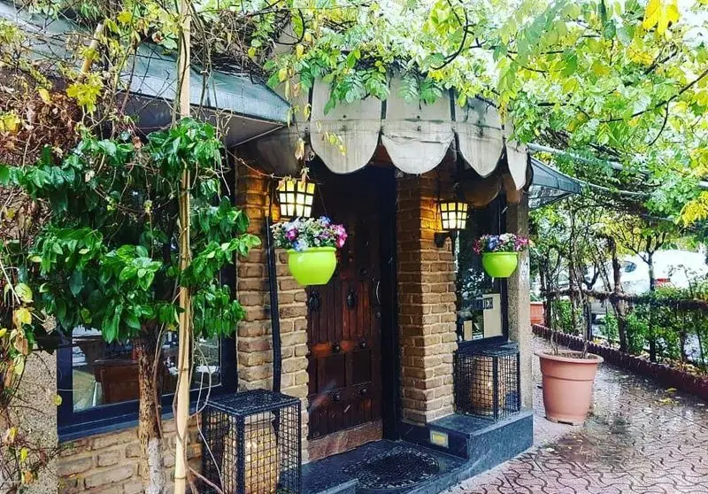 ورودی دنج و زیبای پیتزا دهکده با نمای آجری، درب چوبی، گلدان‌های گل رنگارنگ و پوشش گیاهی سرسبز. محیطی دلنشین برای مشتریان.