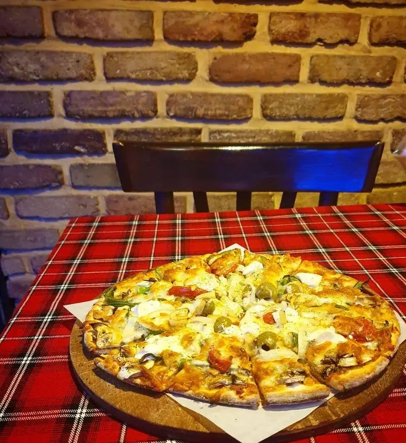 پیتزا دهکده: یک پیتزای بزرگ و تازه با مواد اولیه باکیفیت شامل قارچ، زیتون، فلفل دلمه‌ای و مرغ، روی میز چهارخانه قرمز در فضایی دلنشین.