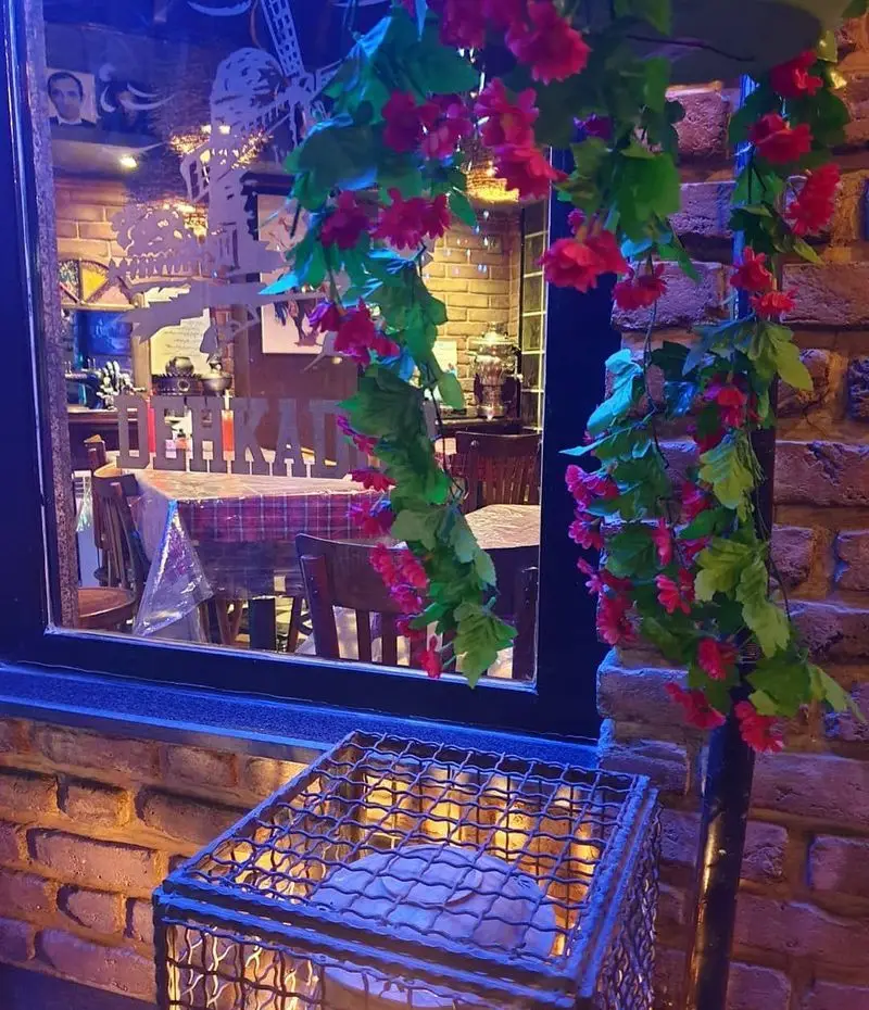 نمای دلنشین پیتزا دهکده: فضای داخلی گرم و دنج با میزهای آماده از پشت پنجره. دکوراسیون آجری بیرون همراه با گل‌های آویزان قرمز.