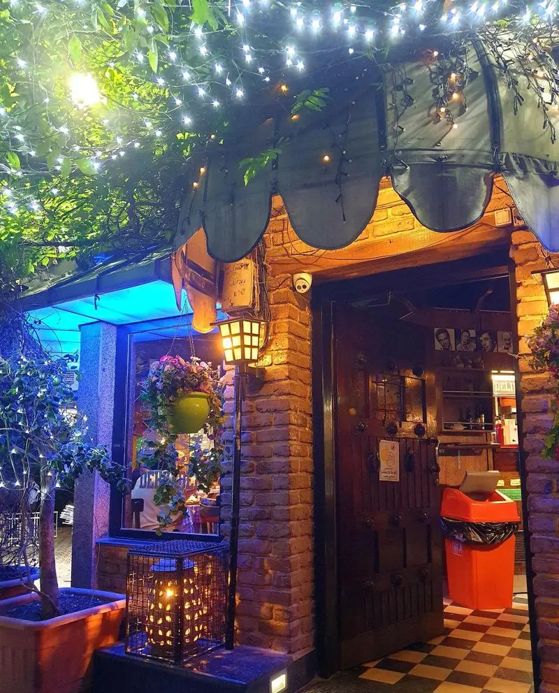 ورودی جذاب پیتزا دهکده در شب با نورپردازی دلنشین ریسه‌ای، آویزهای گل، دیوار آجری و دکوراسیون گرم و دعوت کننده رستوران.