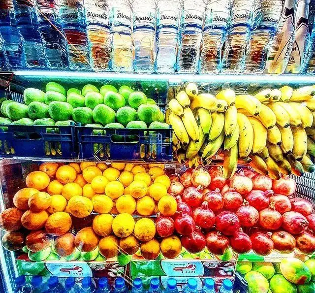 نمایش چیدمان میوه‌های تازه (موز، انار، پرتقال، سیب) و بطری‌های آب در ویتامینه بستنی لئون ترافیک برای آبمیوه طبیعی و سالم.