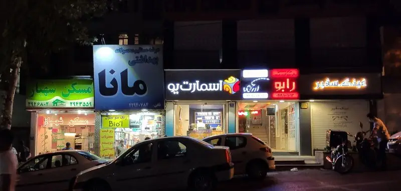 نمای شبانه خیابانی پر جنب‌وجوش، با تابلوهای روشن مغازه‌هایی مانند رستوران آروما، مسکن سهبار، و عینک سفیر، و حضور خودروها در خیابان.