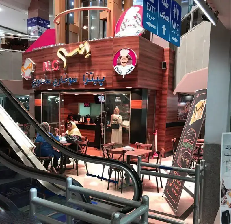 فست فود انسی (NC) در فودکورت مرکز خرید با لوگوی عمو بارنز، ارائه دهنده پیتزا و ساندویچ، دارای میز و صندلی برای مشتریان.