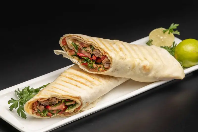 ساندویچ کباب گریل خوشمزه کافه گریل ببک، شامل گوشت لذیذ، سبزیجات تازه و گوجه، با نان تست شده و تزئین لیمو و جعفری.