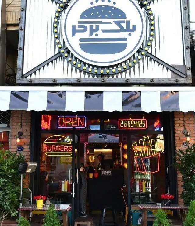 نمای بیرونی فست فود بلک برگر با تابلوی بزرگ لوگو، ورودی روشن با نئون‌های "OPEN" و "BURGERS" در محیطی دلپذیر.