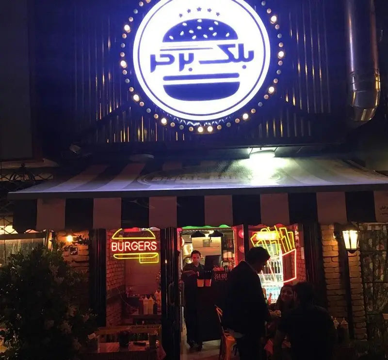 تصویری از نمای شب فست فود بلک برگر با تابلوی اصلی نورانی و لوگوی برگر، تابلوهای نئونی 'برگرز' و سیب زمینی سرخ کرده در محیطی پر جنب و جوش.