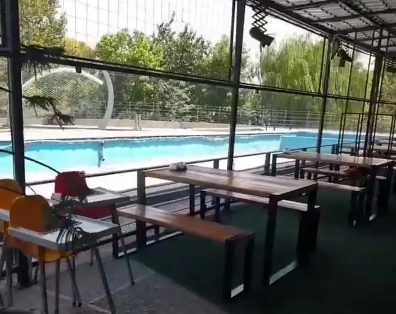 تصویری از فضای دلنشین و باز رستوران کی اف سی سوگود ولیعصر، شامل میز و نیمکت‌های چوبی کنار استخر آبی با نمای طبیعت