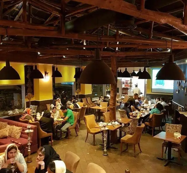 فضای داخلی کافه رستوران سنتی دفینه با دکوراسیون چوبی گرم و صمیمی. مشتریان در حال صرف غذا، نوشیدنی و قلیان در محیطی آرام.