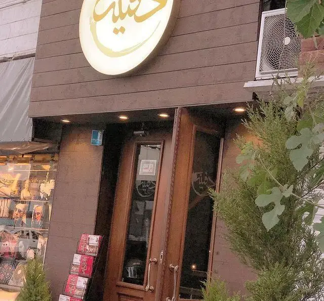 ورودی سرای سنتی دفینه با تابلوی نوری طلایی دفینه، درب‌های چوبی زیبا، و قفسه مجلات، محیطی گرم و دلنشین در انتظار شماست.