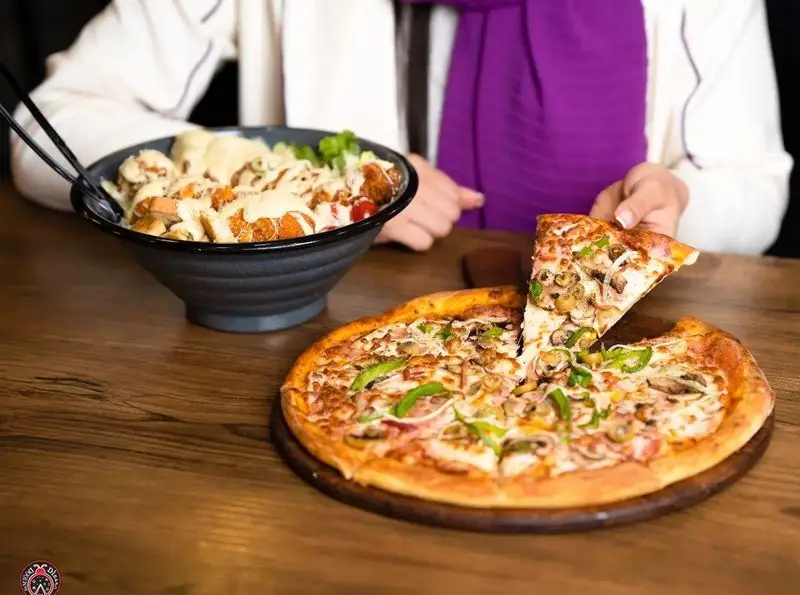 مشتری در حال برداشتن برش پیتزا قارچ و فلفل در کنار کاسه بزرگ سالاد مرغ سوخاری با سس در کافه رستوران ایتالیایی دیبارو.