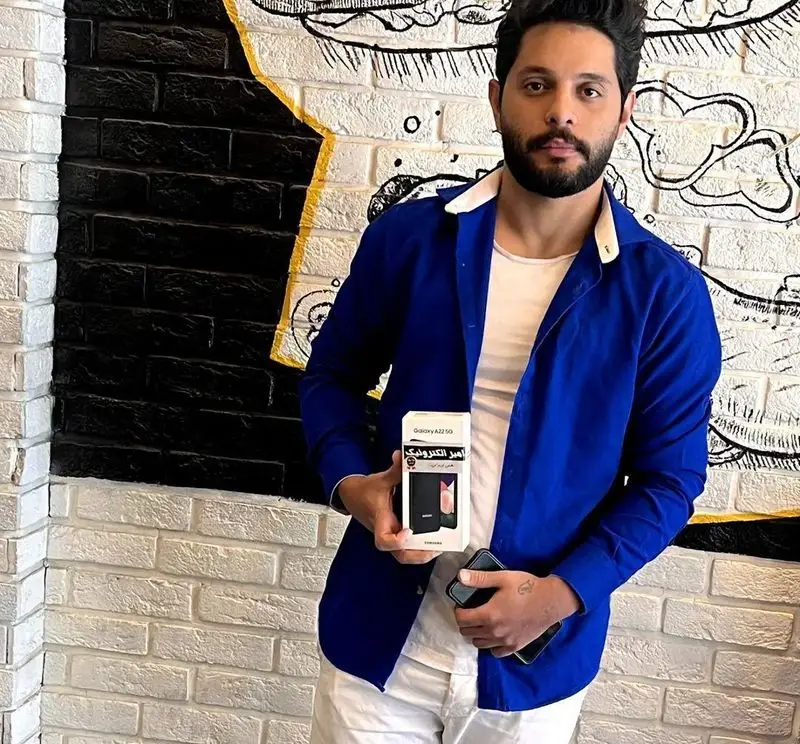 مردی خوش‌تیپ با پیراهن آبی، گوشی جدید سامسونگ گلکسی A22 5G را در دست دارد. محیط دلنشین کافه رستوران ایتالیایی دیبارو.