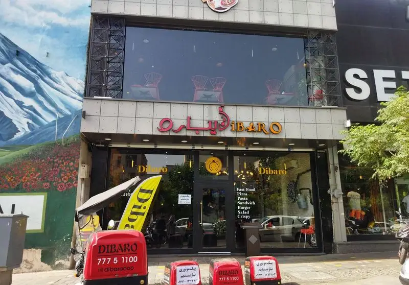 نمای بیرونی کافه رستوران ایتالیایی دیبارو Dibaro، شامل تابلوی بزرگ، ویترین شیشه‌ای و صندوق‌های قرمز رنگ تحویل غذا. منو شامل پیتزا، پاستا، برگر و فست فود.