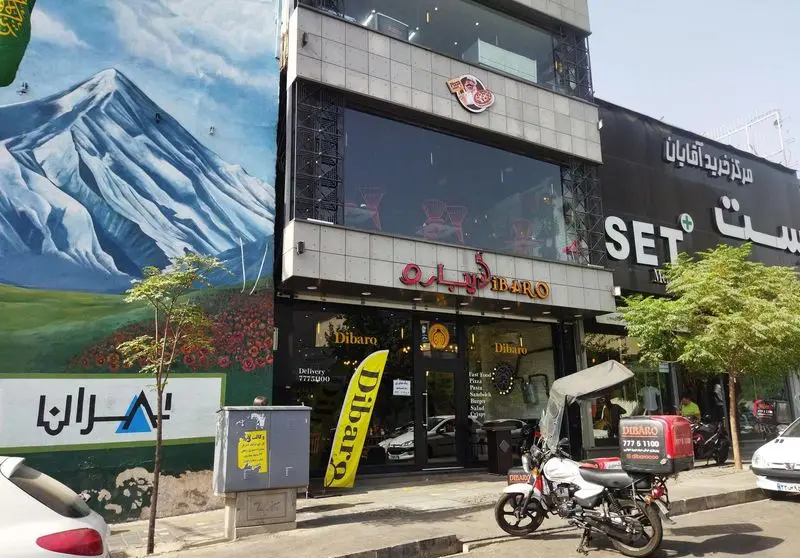 نمای بیرونی کافه رستوران ایتالیایی دیبارو با تابلوی روشن، پرچم زرد و موتور دلیوری، ارائه دهنده بهترین فست فود و پیتزا در تهران.