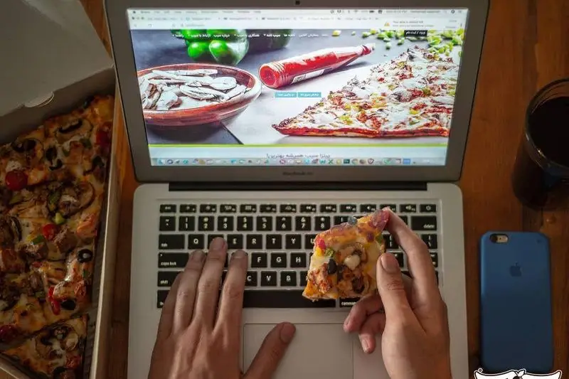 در حال سفارش آنلاین پیتزا از سایت پیتزا سیب 360 و لذت بردن از برش‌های پیتزای گرم و متنوع همراه با نوشیدنی خنک در کنار لپ‌تاپ.