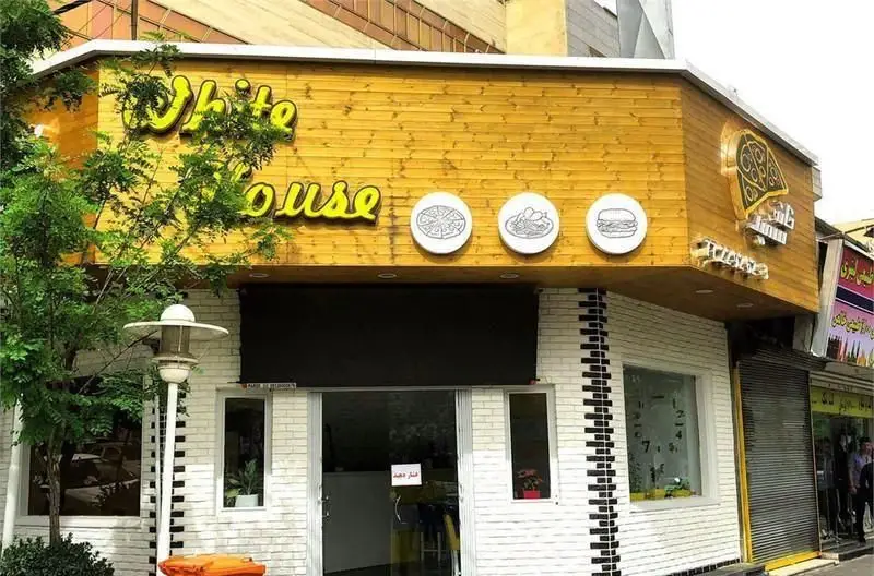 تصویری از نمای خارجی فست فود خانه سفید با تابلوی بزرگ زردرنگ، دیوار آجری سفید و نمادهای پیتزا، پاستا و برگر.