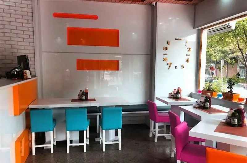 دکوراسیون شیک و رنگارنگ فست فود خانه سفید شامل میزهای سفید، صندلی‌های فیروزه‌ای و صورتی، دیوارهای مدرن و پنجره رو به فضای سبز.