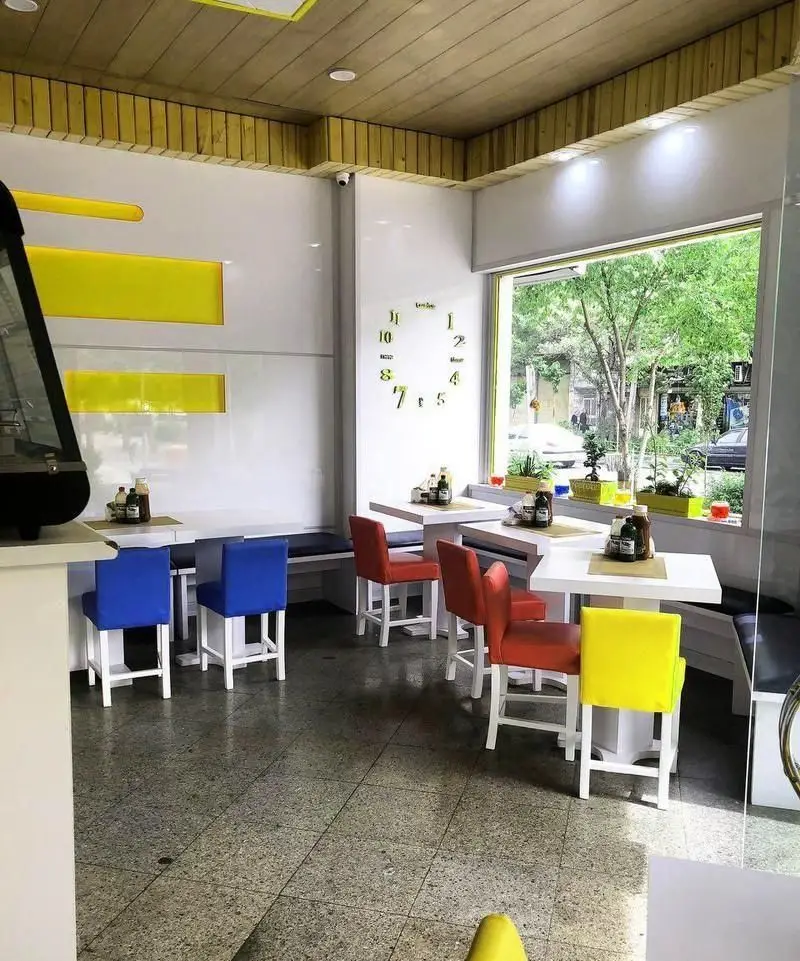 نمایی از فضای داخلی فست فود خانه سفید با دکوراسیون مدرن، دیوارهای سفید، سقف چوبی و میز و صندلی‌های رنگارنگ آبی، قرمز و زرد.
