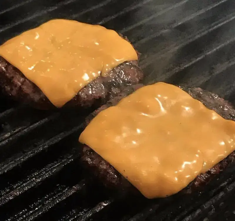 تصویری جذاب از دو چیزبرگر آبدار و لذیذ با پنیر چدار ذوب شده روی گریل. طعم اصیل در برگر جانیس برگر.