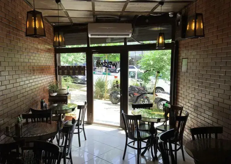 نمایی از محیط داخلی دنج و شیک برگر جانیس برگر با طراحی صنعتی شامل دیوارهای آجری، میزهای شیشه‌ای و صندلی‌های مشکی.