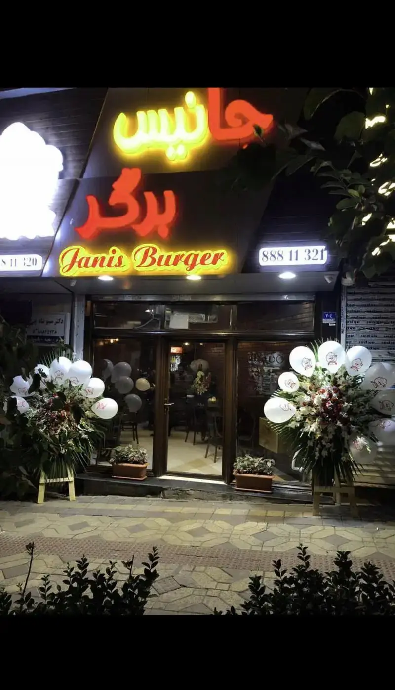 ورودی جذاب فست فود جانیس برگر در شب با تابلوهای نئونی درخشان، گل‌آرایی زیبا و بادکنک‌ها. بهترین برگرها را اینجا امتحان کنید.