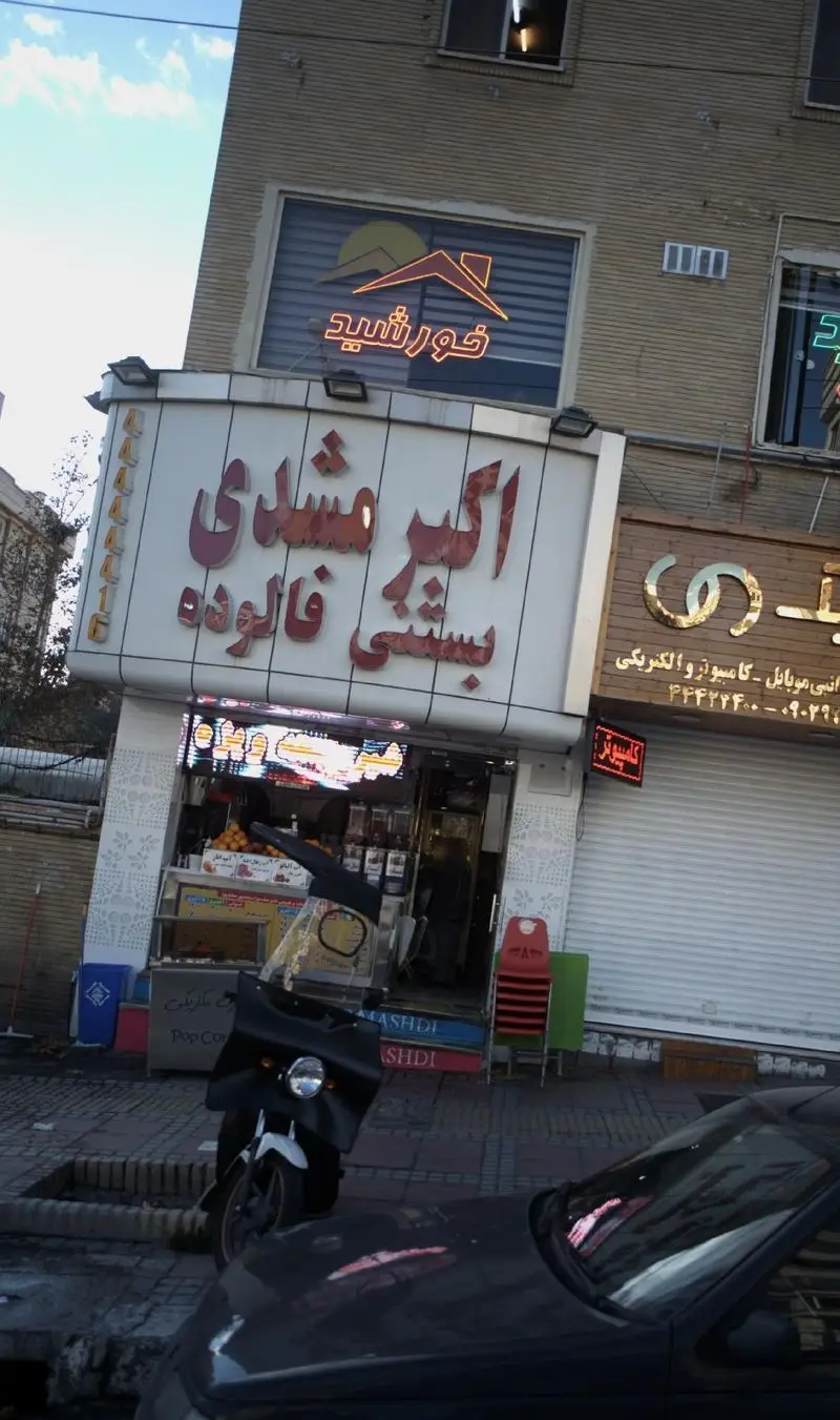مغازه بستنی و فالوده اکبر مشهدی با تابلوی بزرگ و نئون در خیابان، نمایش دهنده محیط شلوغ شهری و کسب و کارهای محلی ایران.