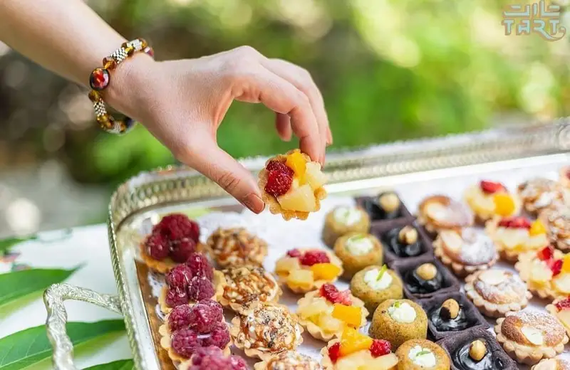 دست در حال برداشتن مینی تارت میوه ای خوشرنگ از سینی پر از مینی تارت های متنوع و شیرینی های لذیذ تارت برای پذیرایی.